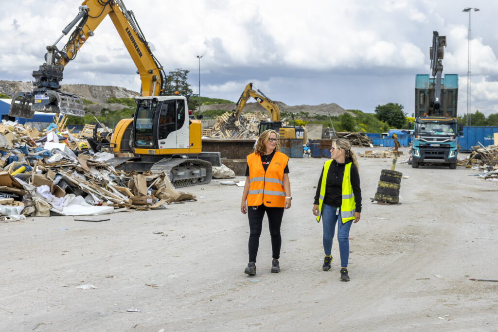 Två personer går på avfallsanläggning med maskiner i bakgrunden